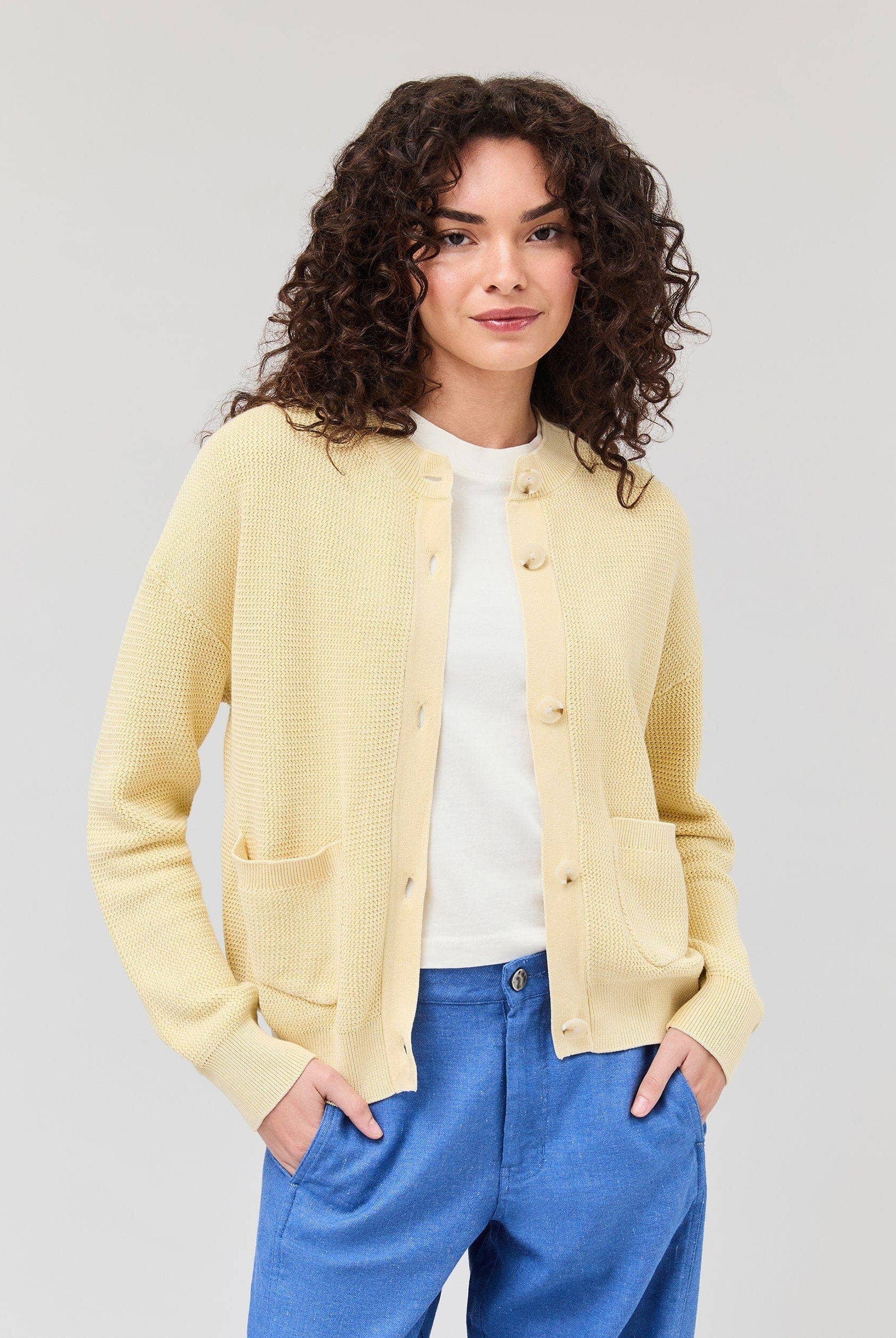 Person wearing a light yellow cardigan over a white shirt with blue pants on a plain background
